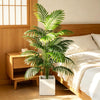An artificial areca palm plant in a white pot sits on a wooden nightstand next to a bed.