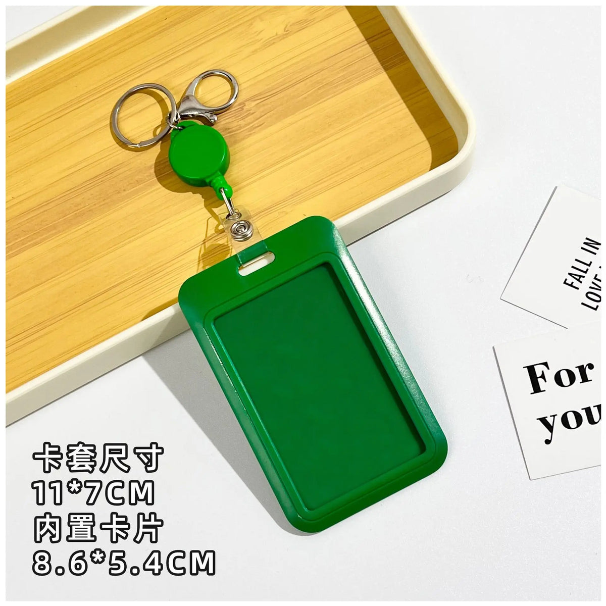 A green retractable ID badge holder with a clip is placed on a wooden tray next to a keychain.