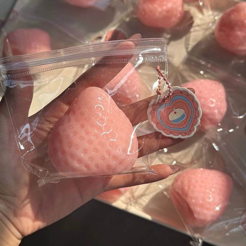 A hand holds a pink strawberry squeeze fidget toy with a decorative charm.