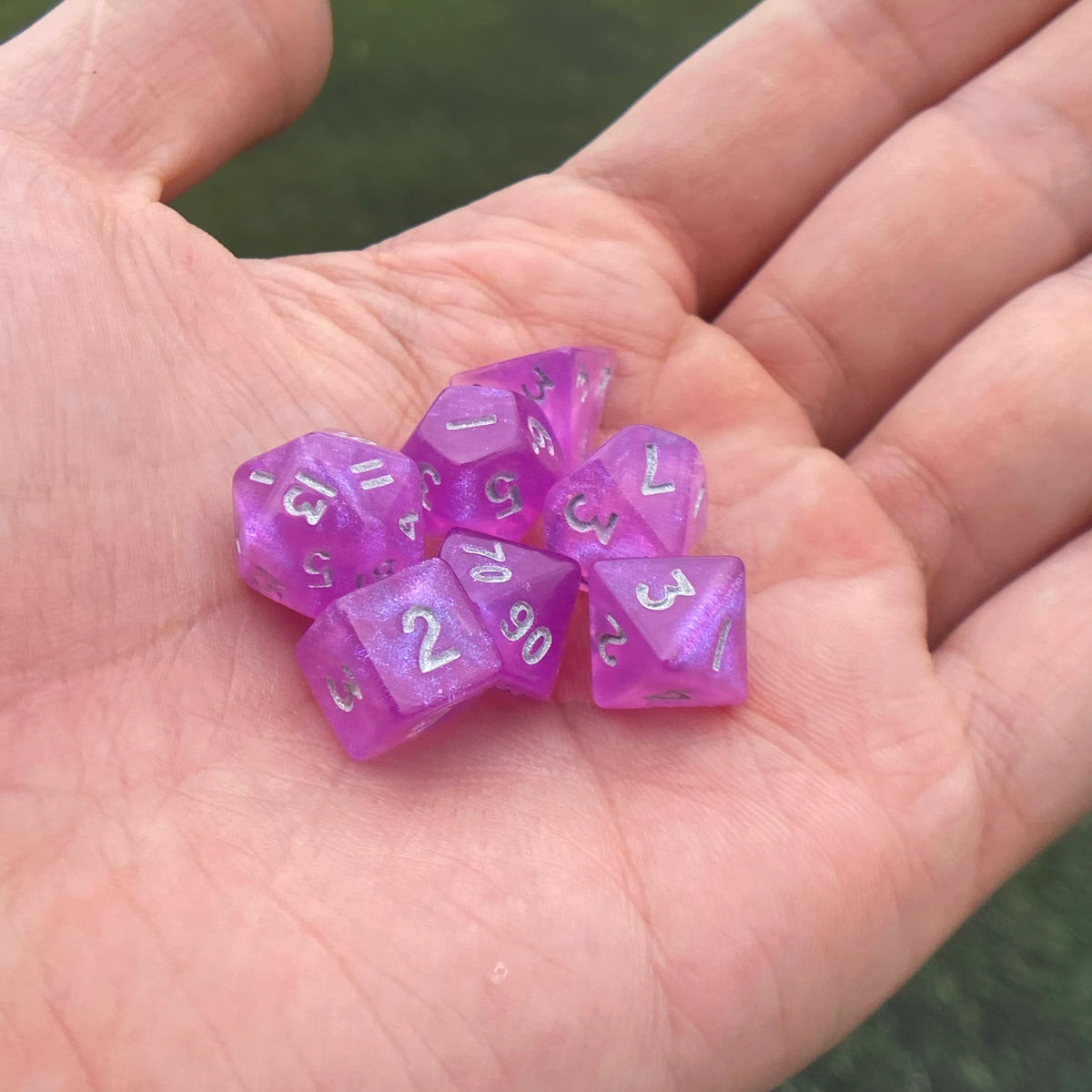 A hand holds a set of seven iridescent purple acrylic polyhedral dice.