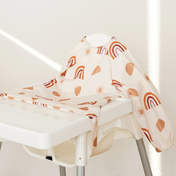 A light beige feeding bib with a rainbow print is displayed on a white stool.
