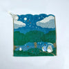 A teal knitted plush toy depicting a night sky scene with clouds and a small owl character.