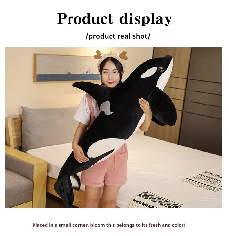A woman is posing with a large black and white orca whale plush toy on a bed.