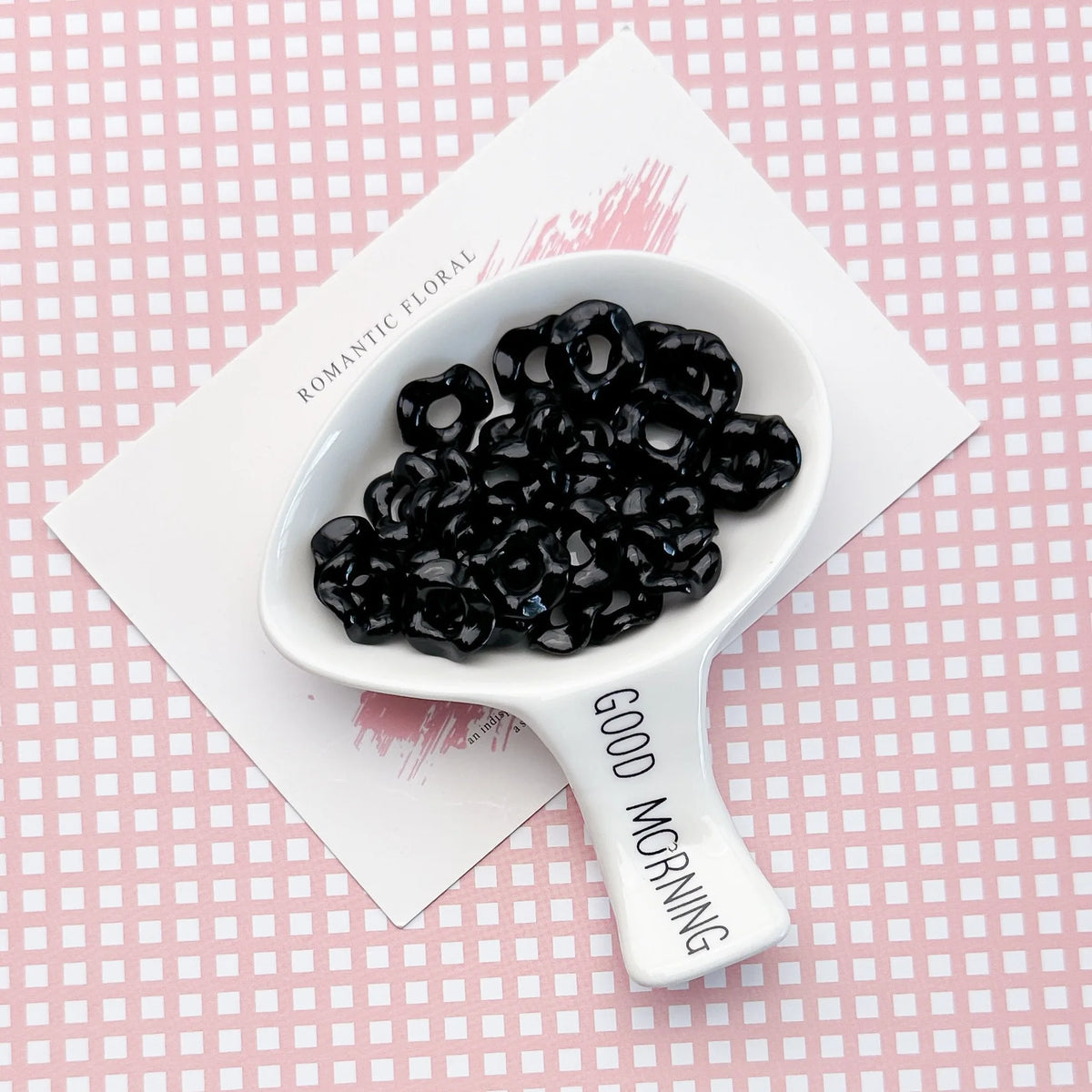 A white spoon holds black acrylic beads with a floral edge on a pink checkered background.