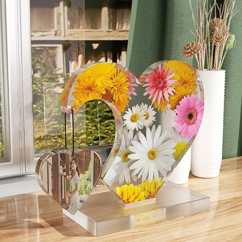 A clear resin heart photo frame mold with floral designs sits on a glass stand next to a potted plant.