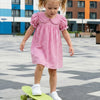 A young girl wearing a pink gingham dress is riding a green skateboard on a paved area.