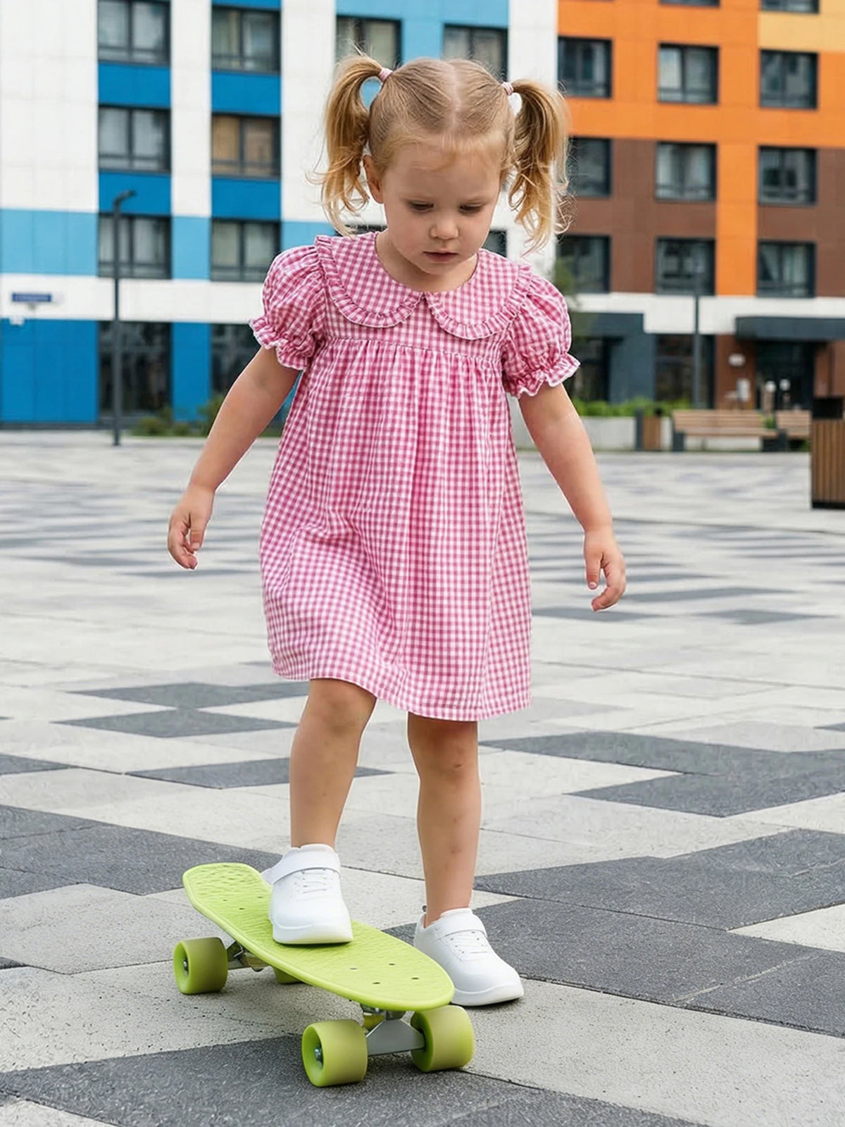 A young girl wearing a pink gingham dress is riding a green skateboard on a paved area.