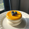 A round, layered dessert featuring a yellow soufflé top and white cream base, garnished with blueberries.