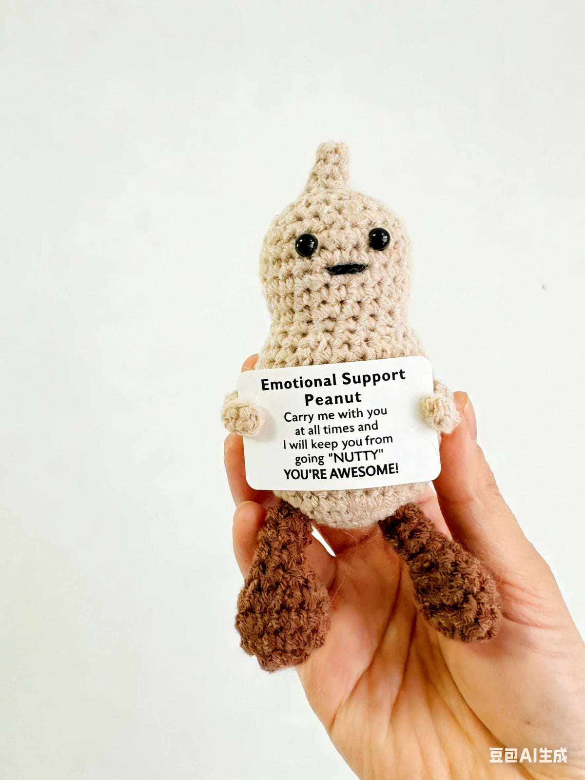 A hand holds a handmade crochet animal plush doll with a sign that reads Emotional Support Peanut