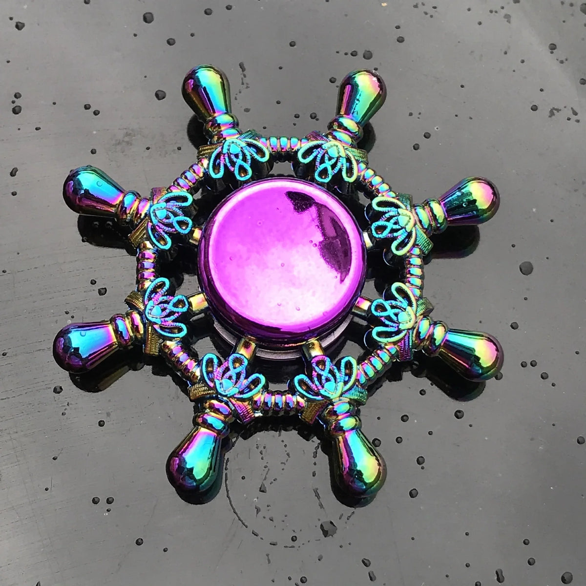 A colorful metal fidget spinner with a purple center and ornate metal arms resting on a dark, wet surface.