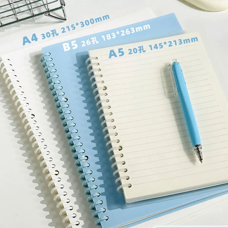 A stack of spiral notebooks in various sizes and colors, including A4, B5, and A5, with a blue pen resting on top.