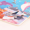 A wooden tangram puzzle set featuring various animal and vehicle shapes in pink, purple, and gray on a light blue background.