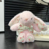 A pink bunny plush toy with a floral bow charm sitting on a white surface.