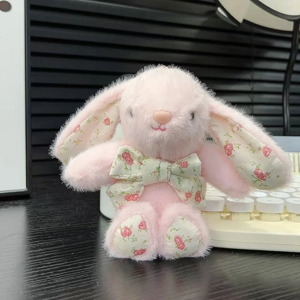 A pink bunny plush toy with a floral bow charm sitting on a white surface.