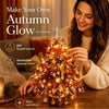 A woman is decorating a DIY fall tree kit with lights and ornaments on a wooden table.