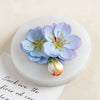 A light blue purple 3D flower silicone mold with green stamens rests on a white circular base.