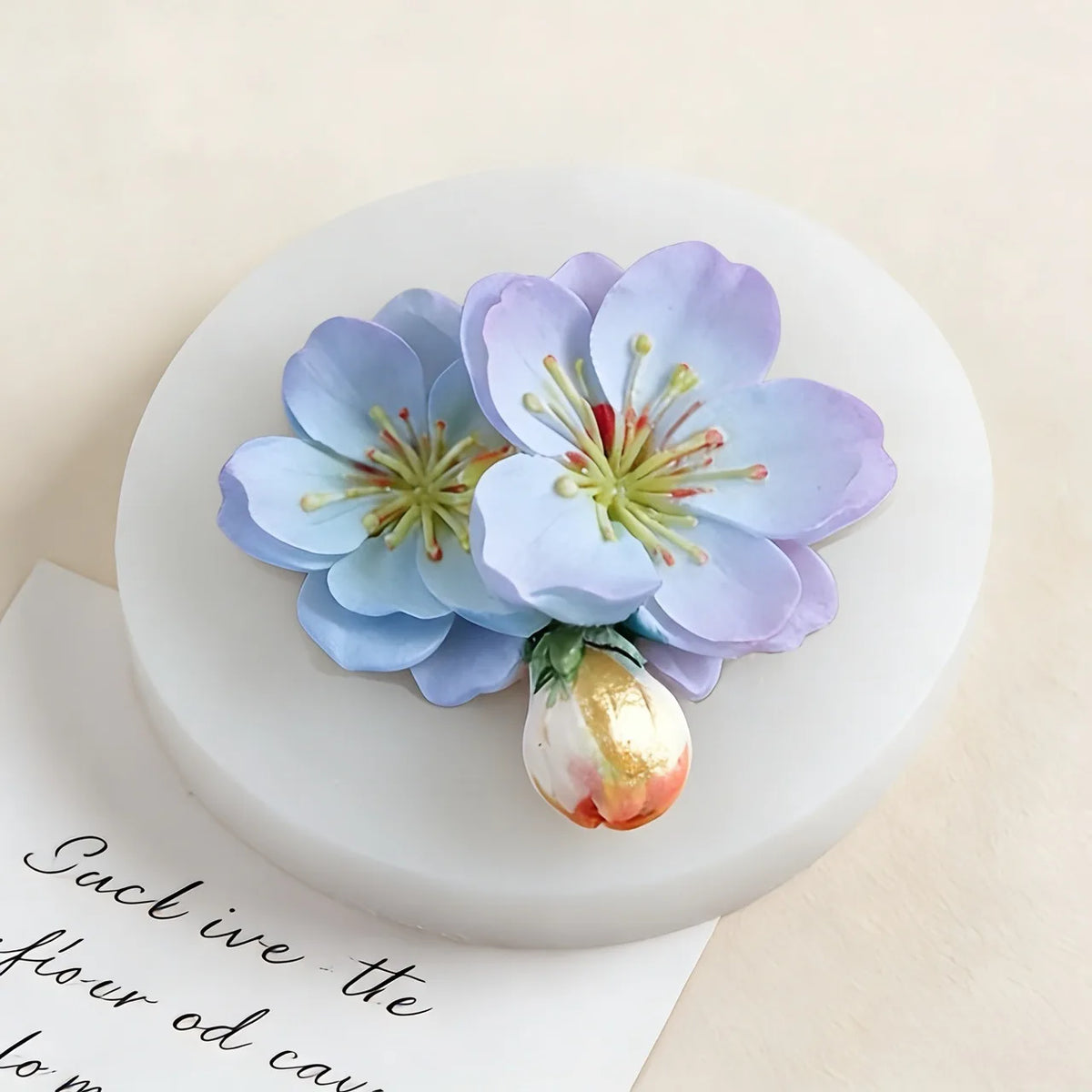 A light blue purple 3D flower silicone mold with green stamens rests on a white circular base.