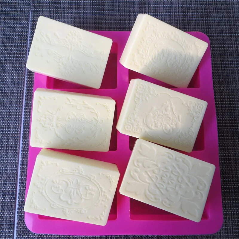 Six square silicone molds with floral patterns for DIY soap making arranged in a pink tray.