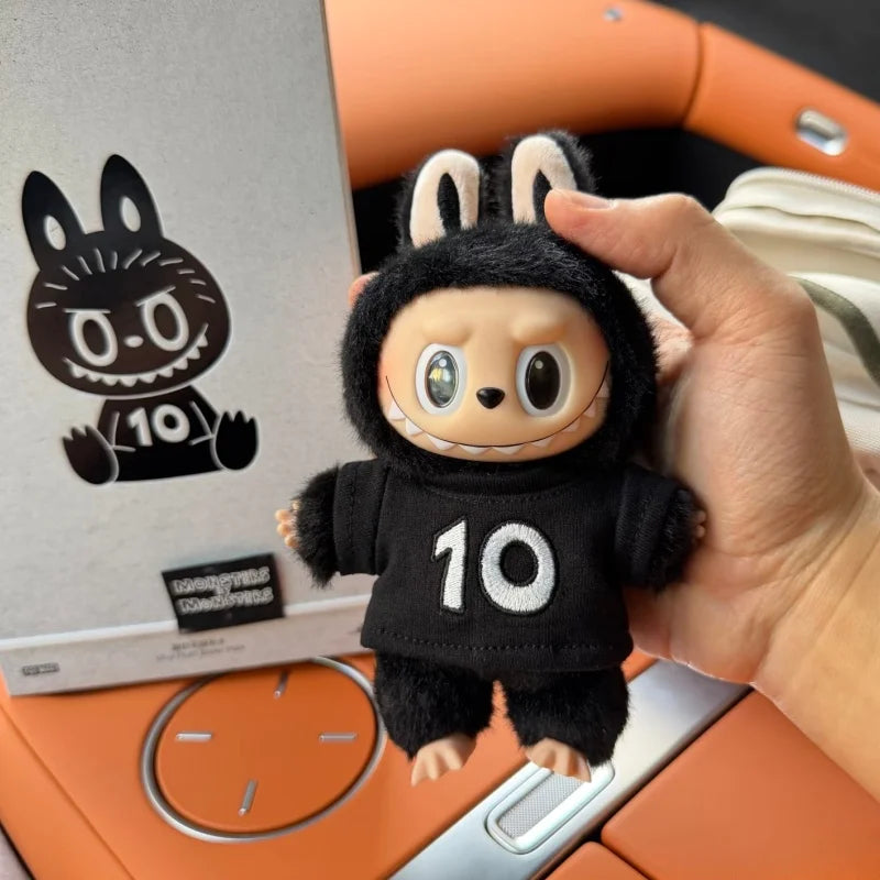 A hand holds a black plush toy resembling a rabbit with large eyes and a white toothy grin, set against an orange background.