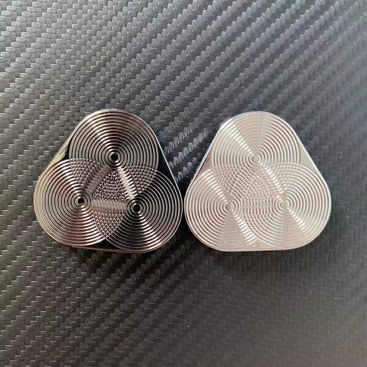 Two silver metal magnetic fidget sliders resting on a dark carbon fiber surface.