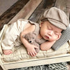 A newborn baby is sleeping wearing a beige knit romper and a matching newsboy cap, resting on a light-colored fabric.