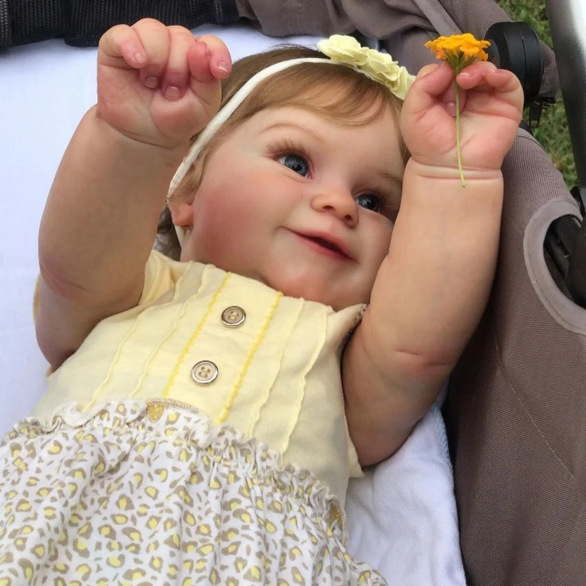 A close-up of a lifelike 48cm soft vinyl baby doll with 3D skin detail, wearing a yellow top and holding a small orange flowe