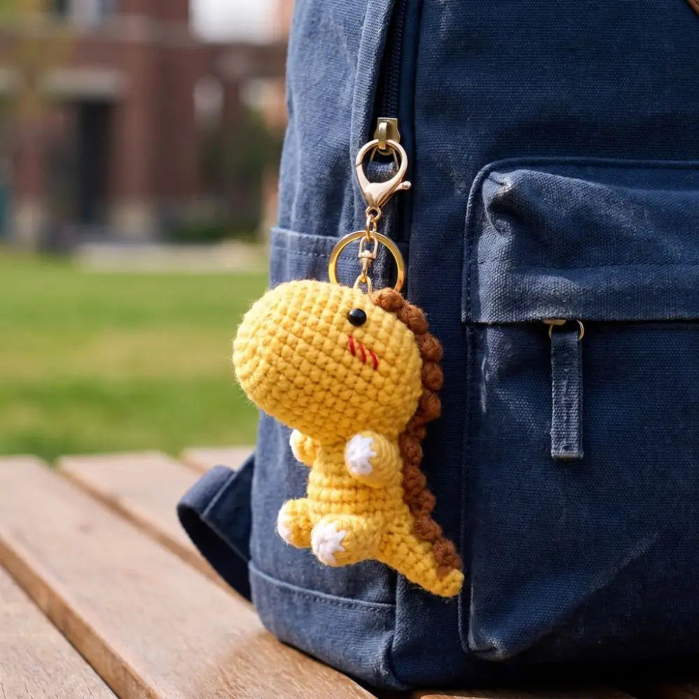 A yellow crochet dinosaur keychain with brown hair is attached to a dark blue fabric backpack.