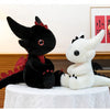 A black Toothless dragon plush toy sitting next to a white unicorn plush toy on a white draped surface.