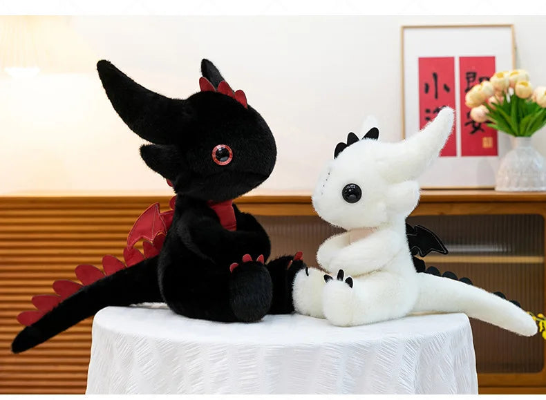 A black Toothless dragon plush toy sitting next to a white unicorn plush toy on a white draped surface.