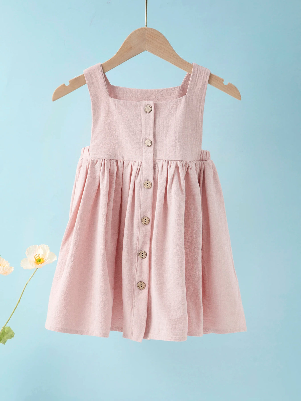 A pink linen jumper dress for a toddler girl hanging on a wooden hanger against a light blue background.