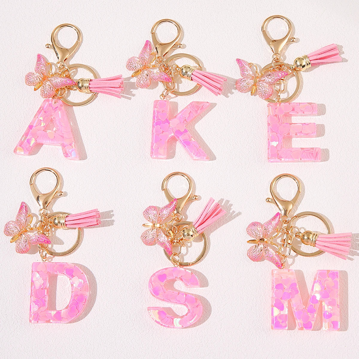A set of initial heart keychains featuring pink glitter acrylic letters and gold hardware with pink feather butterfly charms.