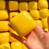 A hand holds a bright yellow sticky cube slow-rising squishy stress relief toy with small red dots.