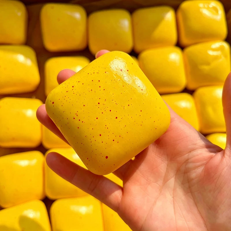 A hand holds a bright yellow sticky cube slow-rising squishy stress relief toy with small red dots.