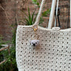A close-up of a light beige crocheted animal keychain, possibly a rabbit or chick, hanging from a natural fiber strap.