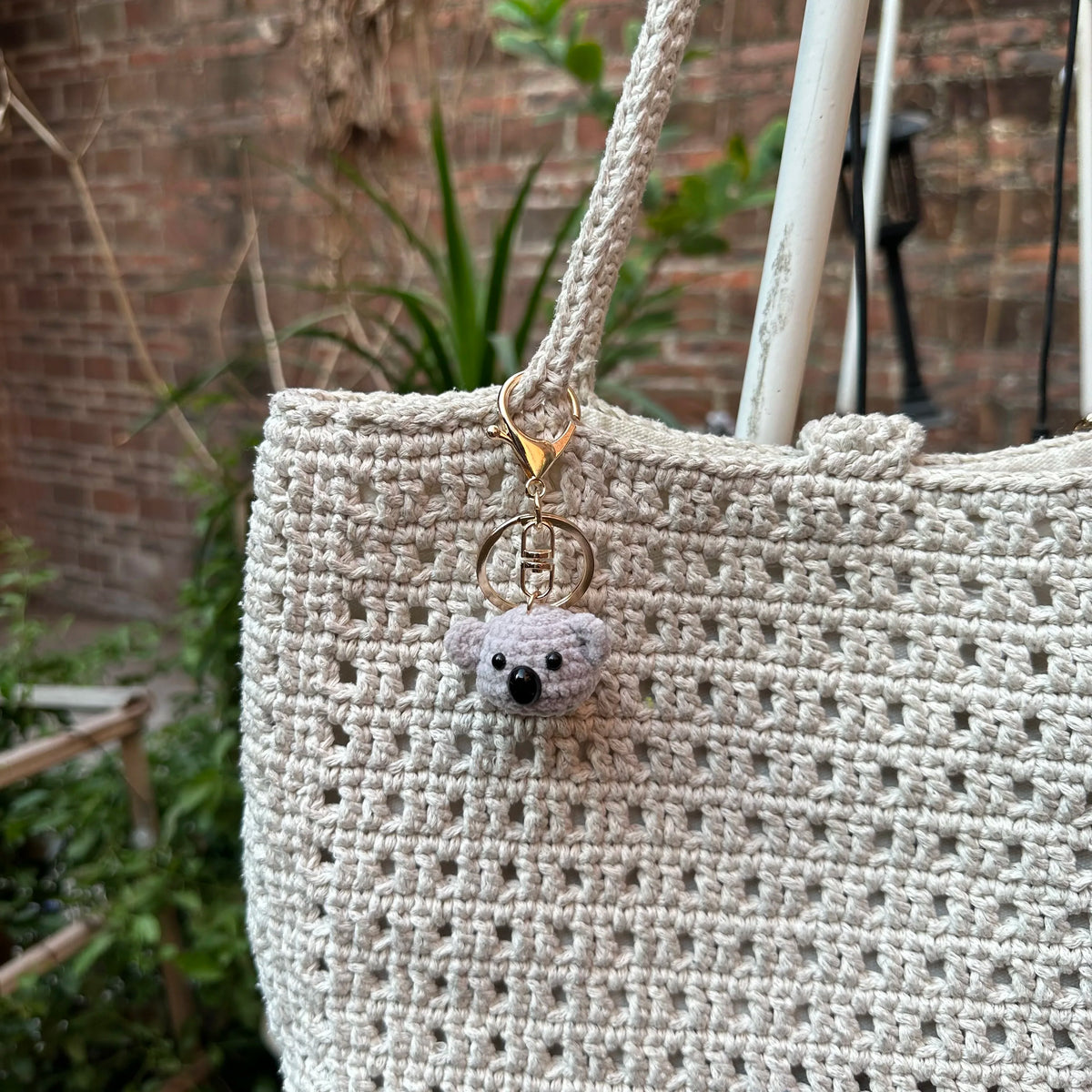 A close-up of a light beige crocheted animal keychain, possibly a rabbit or chick, hanging from a natural fiber strap.
