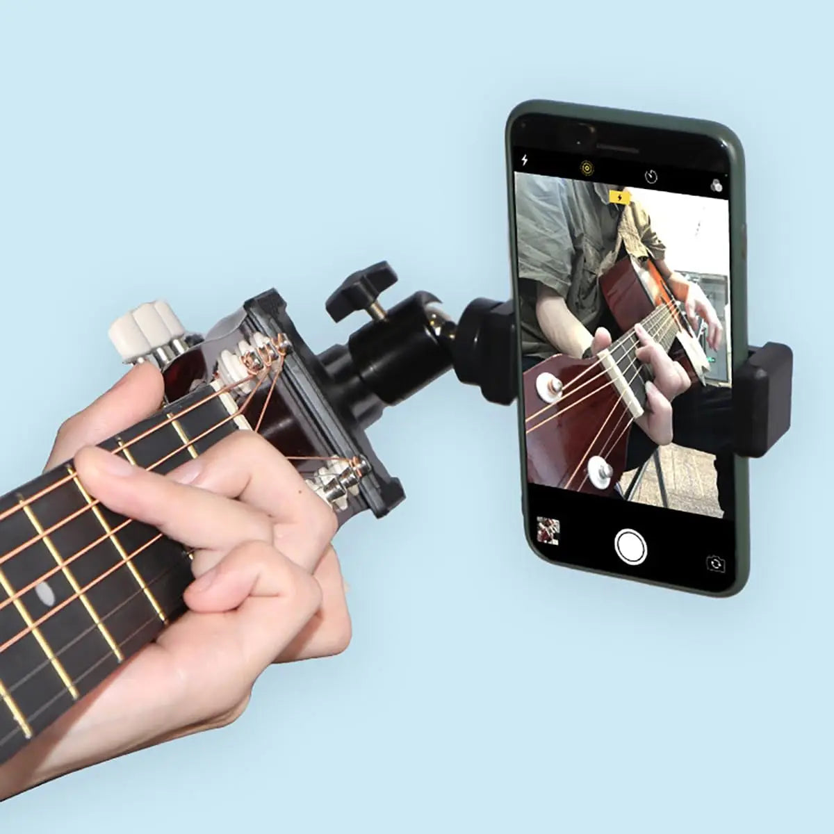 A hand plays a guitar while it is mounted on a phone for live streaming or recording.