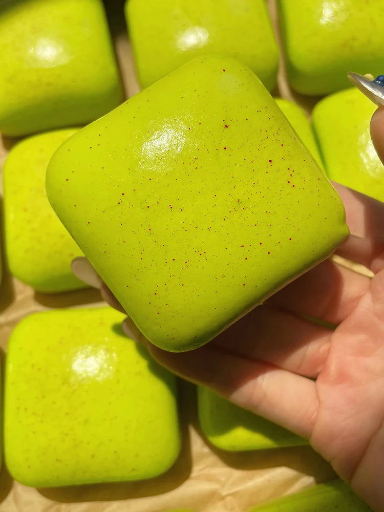 Eine leuchtend grüne, quetschbare Simulation von Brot als Stressball-Spielzeug wird in einer Hand gehalten, mit ähnlichen Spi