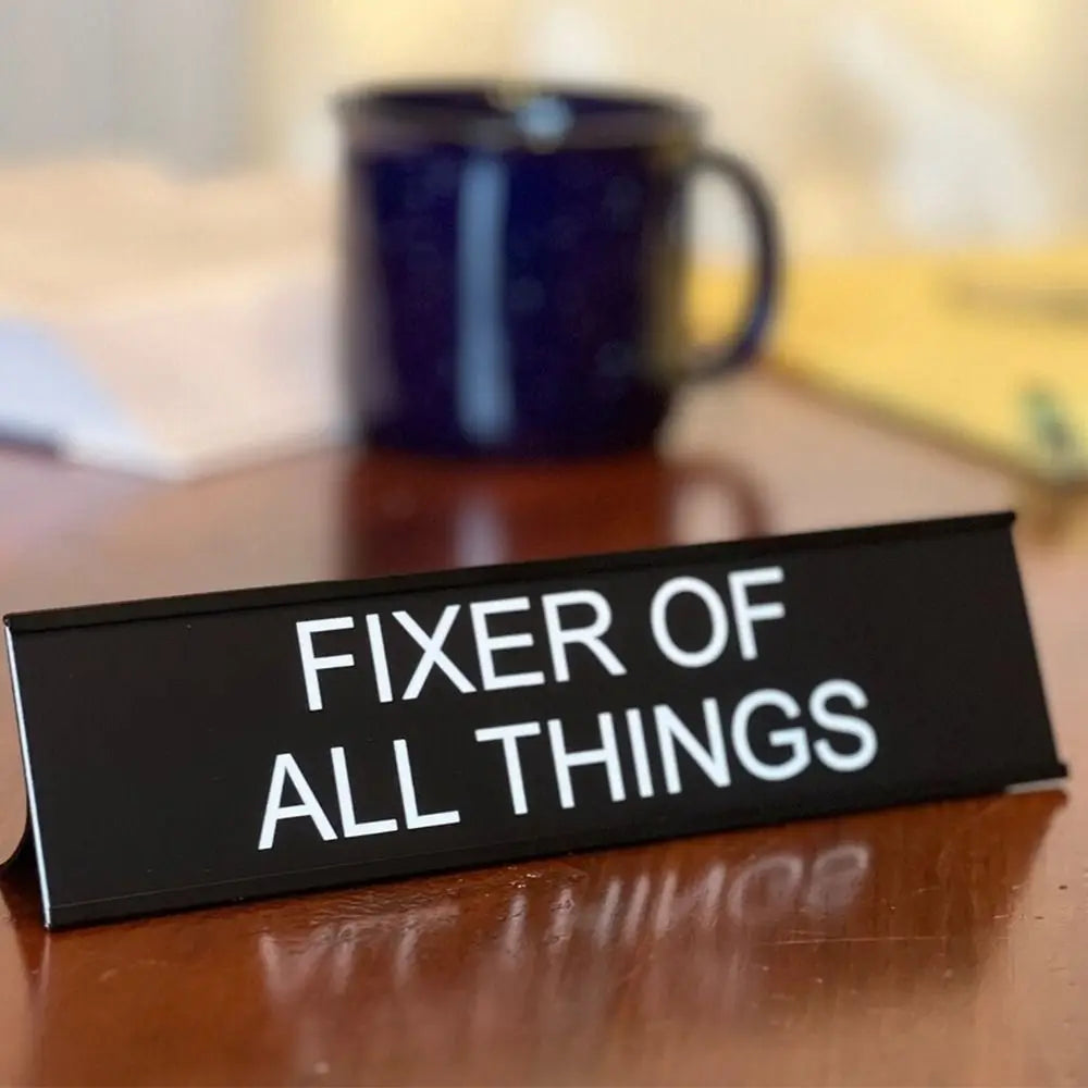 A black desk sign with white text that reads Fixer of All Things, resting on a wooden surface.
