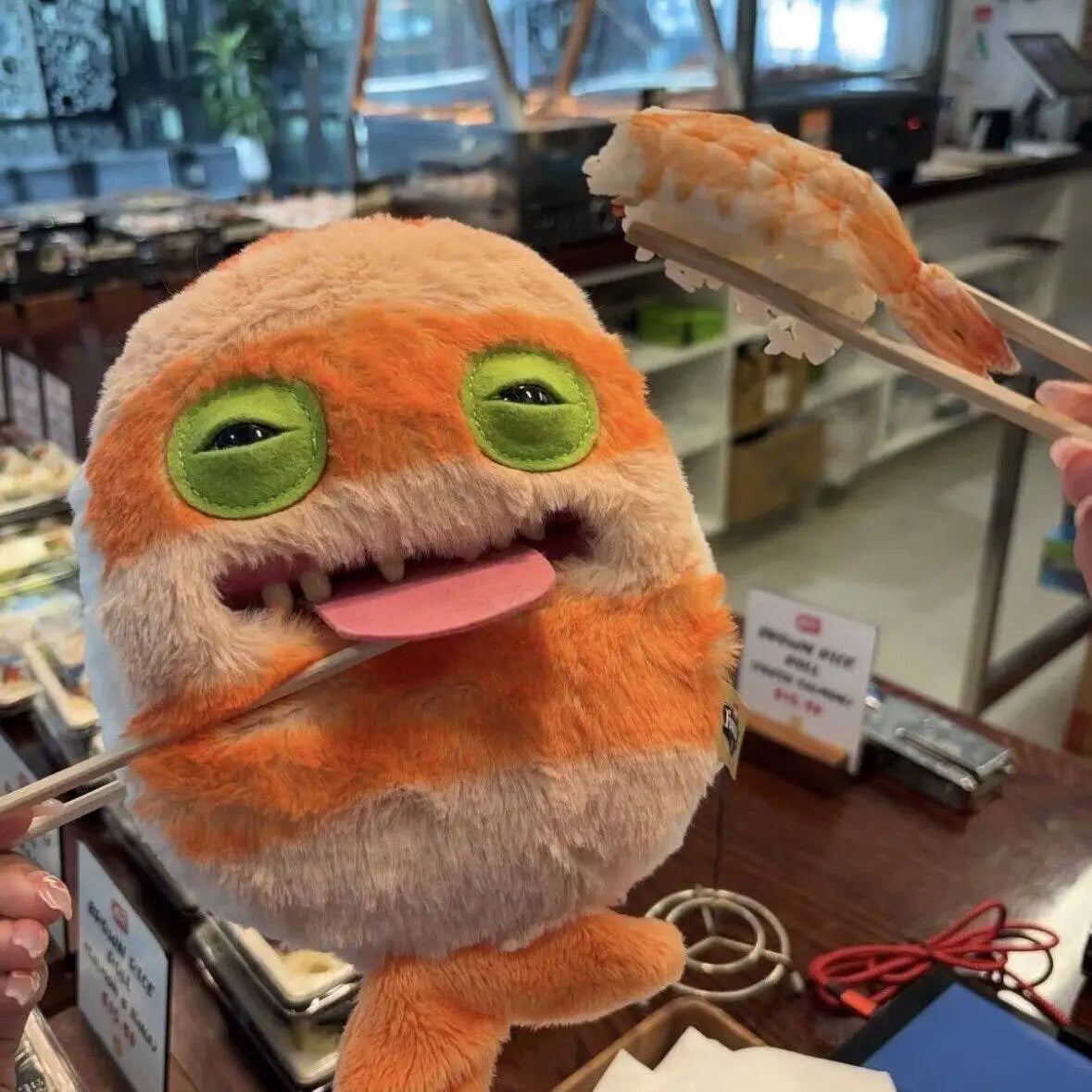 An orange and white monster plush toy with large green eyes is being held by chopsticks.