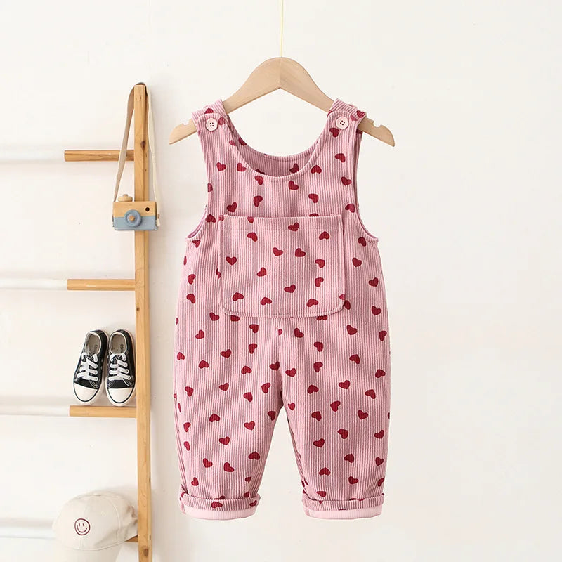 Pink corduroy toddler overalls with a pattern of red hearts hanging on a wooden hanger.