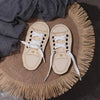 A pair of light tan wooden lacing shoes resting on a woven mat with fringe, next to a gray cloth.