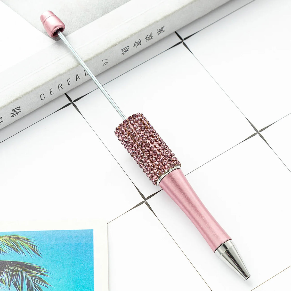 A pink beaded ballpoint pen craft kit with a silver needle is displayed on a white surface next to a book.
