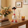 A white goose figurine wearing a black hat, glasses, and necklaces displayed on a wooden table.
