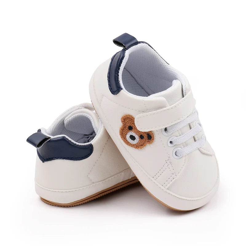 A pair of white baby sneakers with navy blue accents and a brown bear design for first walkers.