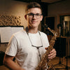A young man holds a gold saxophone with a strap, wearing glasses and a white t-shirt.