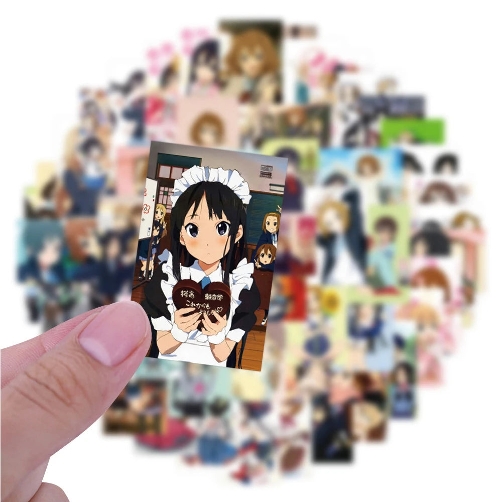 A hand holds a square sticker featuring a cute guitar girl character from K-ON! anime as part of a larger decal pack.