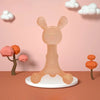 A light peach silicone teether shaped like a bunny stands on a white plate against a pink background.