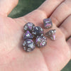 A hand holds a set of seven iridescent acrylic polyhedral dice with a velvet pouch.
