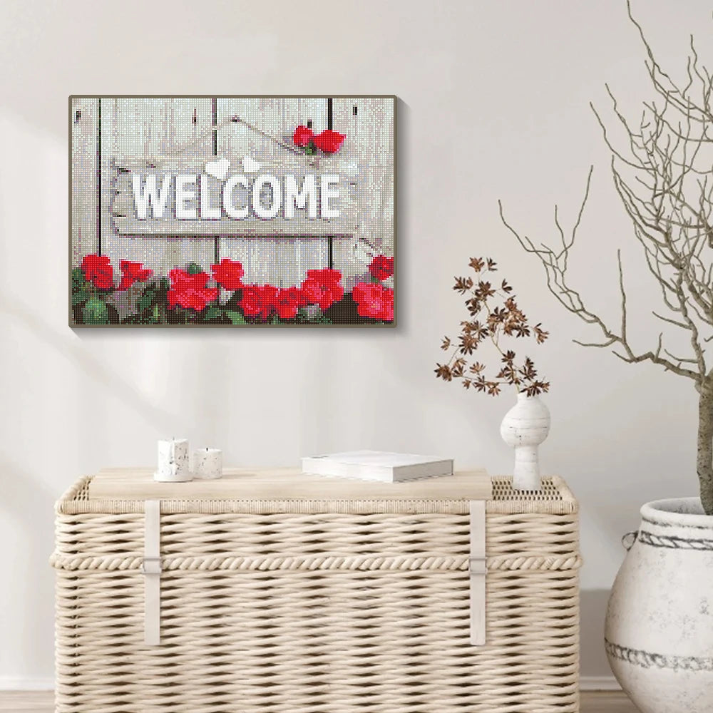 A framed diamond painting featuring the words Welcome on a wooden sign surrounded by red roses and greenery.