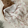A soft white muslin swaddle blanket with a delicate floral print is draped next to a brown teddy bear.